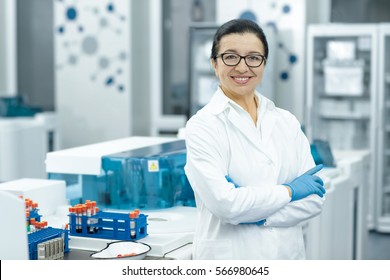 Senior specialist. Cheerful elderly female scientist smiling happily with her arms crossed posing at her lab colleagues on the background teamwork profession professor guidance people job copyspace  - Powered by Shutterstock