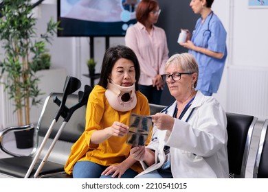 Senior Specialist Consulting Patient With Cervical Neck Collar Looking At Medical Xray Exam During Checkup Visit In Hospital Waiting Room. Asian Woman With Physical Pain And Injury Having Appointment.