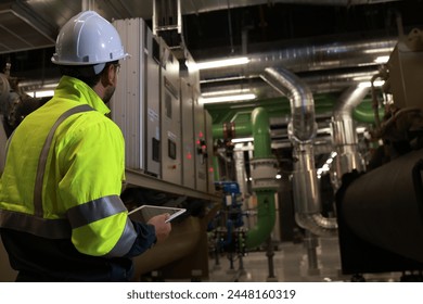 Senior specialist engineer wearing green safety jacket hold digital tablet working preventive maintenance industrial HVAC machinery 