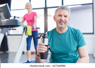 senior sportsman sitting with sport bottle, sportswoman on treadmill behind. senior fitness class concept  