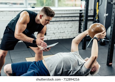 Senior Sportsman Training On On Fitness Ball With Medicine Ball In Gym While Trainer Using Digital Tablet