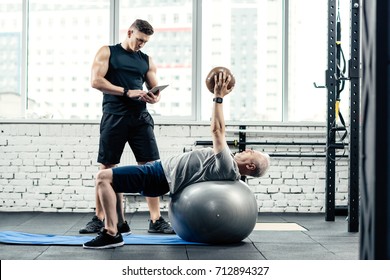 Senior Sportsman Training On On Fitness Ball With Medicine Ball In Gym While Trainer Using Digital Tablet