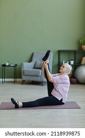 Senior Sportswoman In Beige T-shirt And Black Leggins Holding And Raising Stretched Leg While Exercising On Yoga Mat