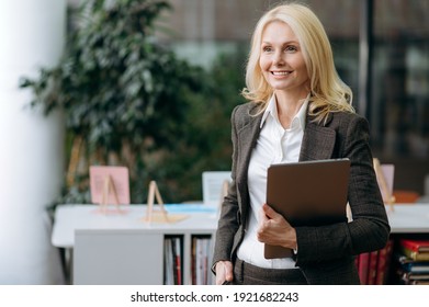 Senior successful female employee standing in modern office, hold a laptop in arms. Happy blonde-hair business lady in elegant stylish suit looks away and smiling - Powered by Shutterstock