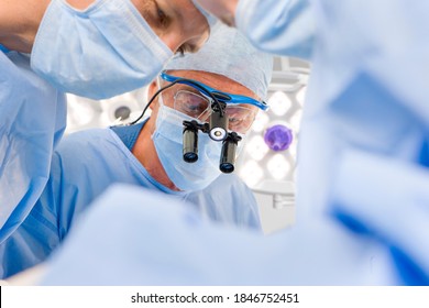 A senior surgeon performing an operation with his teammates in the operating room while wearing surgical binoculars - Powered by Shutterstock