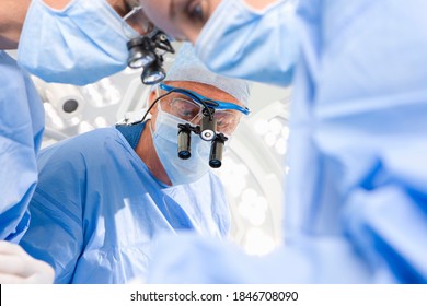 A Senior Surgeon In Selective Focus Looks Down At The Operation Table The Surgical Loupe During A Serious Operation With His Coworkers In The Foreground