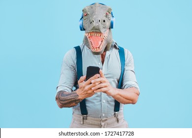 Senior tattooed man with t-rex mask using smartphone while listening music during Coronavirus time - Crazy man wearing funny mask for Covid-19 prevention - Focus on face - Powered by Shutterstock