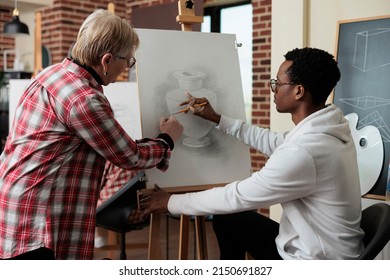 Senior teacher explaining sketching technique to african american student during art lesson in creativity studio  Artist man drawing vase model canvas learning new sketch skill for personal grow