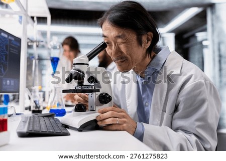 Senior technician operating compound microscope, doing detailed sample observation in controlled laboratory environment. Asian man studies specimen under optical device in biotechnology lab