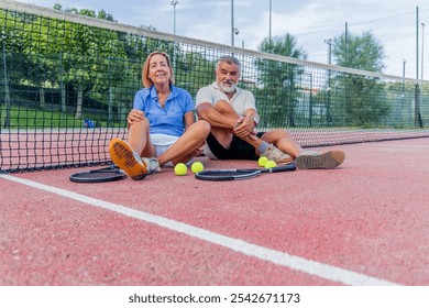 senior tennis couple sits together by the net on the court, holding their rackets and tennis balls, smiling at the camera. Their relaxed and confident expressions show their love for the game - Powered by Shutterstock