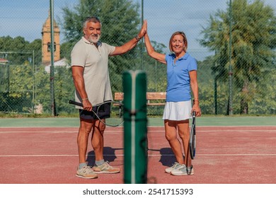 senior tennis couple smiles and shares a high-five across the net while looking at the camera, celebrating their teamwork and enthusiasm. This moment highlights their active lifestyle - Powered by Shutterstock