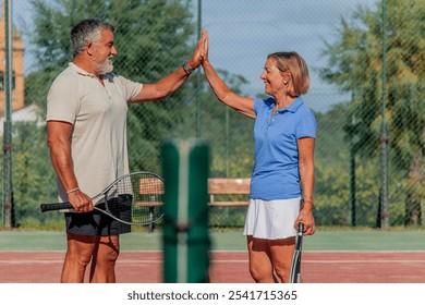 senior tennis player shares a joyful high-five with his wife before their match, with the net positioned between them. This playful gesture captures their connection and enthusiasm for the game - Powered by Shutterstock