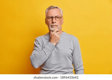 Senior thougthful man holds chin and looks pensively aside makes plannings wears spectacles and casual grey jumper isolated over vivid yellow background. Handsome grandfather considers something - Powered by Shutterstock