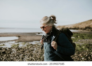 Senior tourist woman walking at the beach. Senior woman on a hike at the beach. Woman walking in nature. Wellness for senior woman, hiking in nature at a cold beach. Tourist by the coast. - Powered by Shutterstock