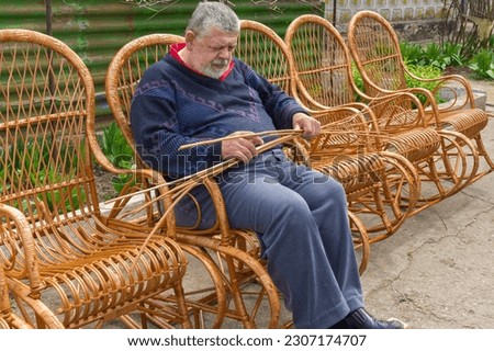 Senior Ukrainian man sorting withe while making whicker rocking chair in summer garden 