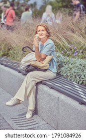 Senior White Attractive Woman About 62 Years Old Is Sitting On The Bench In The Public Park And Eating Ice Cream. There Are Flowers On The Background.