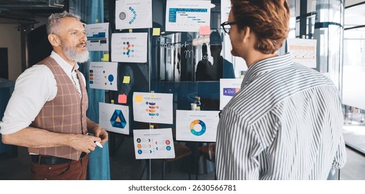 A senior White businessman explains business data to a younger colleague in front of a glass wall filled with colorful charts and infographics in a creative open-plan workspace