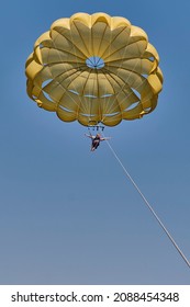 Senior white woman with gray hair is parasailing. She is flying under yellow parachute in blue sky, smiling and opening her arms. Extreme entertainment concept.