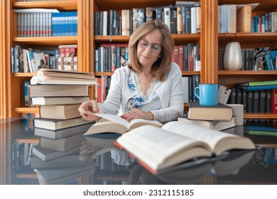 Senior white woman reading a book on a glass table with a bookcase full of book behind her. - Powered by Shutterstock