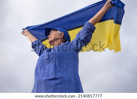 Senior white-haired woman standing outdoors waving the Ukrainian flag looking away  feeling the freedom. No war, stop fights, we want peace