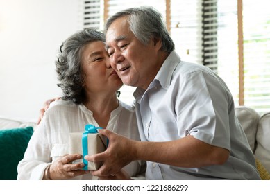 Senior Wife Kissing On Her Husband Cheek. Lovely Asian Older Couple Holding Gift Or Present Box For Birthday Or Christmas Or Wedding Anniversary.