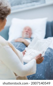 Senior Wife Stands In Front Of The Bed Of Her Sick Husband And Holds An Adult Diaper