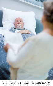 Senior Wife Stands In Front Of The Bed Of Her Sick Husband And Holds An Adult Diaper