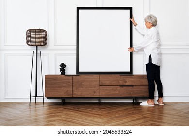 Senior Woman Adjusting A Blank Photo Frame On A Wooden Cabinet In A Japandi Living Room