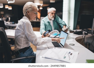 Senior woman analyzes data on a laptop while multitasking with a smartphone, collaborating with a male colleague in a dynamic office setting. - Powered by Shutterstock