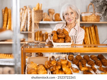 Senior woman baker sell croissants, shows different puff pastry. Authors bakery, fresh pastry. In sales hall near showcase, she demonstrate finished products, lures customers with fragrant pastries - Powered by Shutterstock