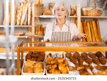 Senior woman baker sell croissants, shows different puff pastry. Authors bakery, fresh pastry. In sales hall near showcase, she demonstrate finished products, lures customers with fragrant pastries - Powered by Shutterstock