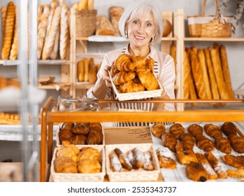 Senior woman baker sell croissants, shows different puff pastry. Authors bakery, fresh pastry. In sales hall near showcase, she demonstrate finished products, lures customers with fragrant pastries - Powered by Shutterstock