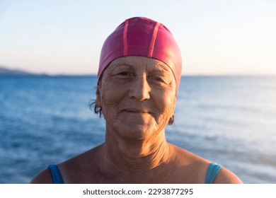 Senior woman at the beach, standing by the sea. Wearing a blue swimsuit and pink swimming cap, ready to swim - Powered by Shutterstock