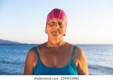 Senior woman at the beach, standing by the sea. Wearing a blue swimsuit and pink swimming cap, ready to swim - Powered by Shutterstock