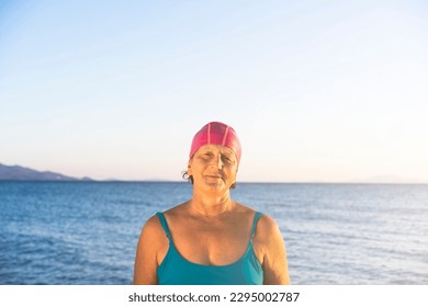 Senior woman at the beach, standing by the sea. Wearing a blue swimsuit and pink swimming cap, ready to swim - Powered by Shutterstock