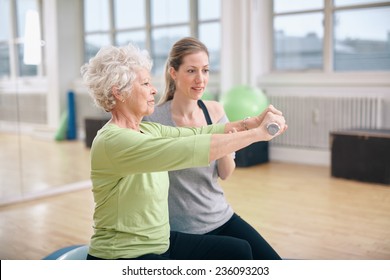 Senior woman being assisted by instructor in lifting dumbbells at gym. Senior woman training in the gym with a personal trainer at rehab. - Powered by Shutterstock