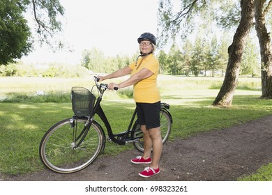 Senior Woman Bicycling In A Park At Sunny Afternoon, Standing Next To A Step Through Bicycle.