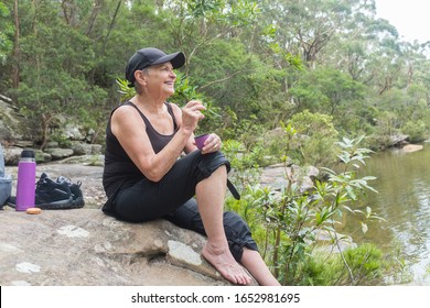 Senior Woman In Black Hiking Clothing Sitting On Rock By Water In Nature Drinking And Eating (selective Focus)