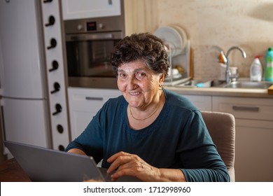 A Senior Woman With Black Short Hair In A Turquoise Shirt Is Looking At The Camera While Working On A Laptop Computer. She Is In The Kitchen And Browsing The Internet For Some New Recipes For Cooking