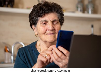 A Senior Woman With Black Short Hair In A Turquoise Shirt Sits In The Kitchen And Uses A Smartphone. She Is Happy And Laughing. The Phone Is Metallic Blue And In Front Of It Is A Laptop Computer.