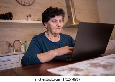 A Senior Woman With Black Short Hair Sits At A Table In The Kitchen And Works On A Laptop Computer. She Is Looking For Some New Cooking Recipes Online And She Is Thoughtful And Very Serious.