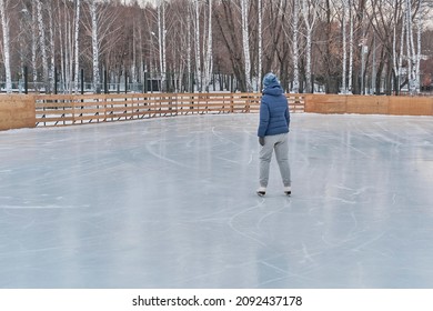 Senior Woman In Blue Jacket And Knitted Hat Skates On A Skating Rink In The Park In Winter. Full Length Rear View. Active Ageing, Body Positive Concept