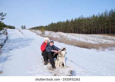 Senior Woman And Boy Sit On Sleigh With Husky Dog In Sledding Sled In Winter Forest 