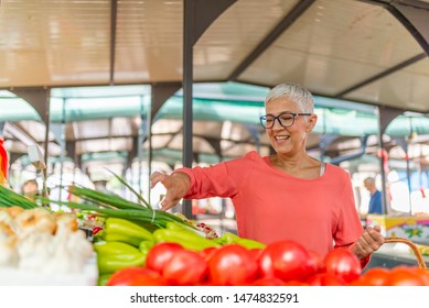 Senior Woman Buying Vegetables At Farmers Market. Woman Picking Fresh Produce At The Market. Senior Woman Shopping At An Outdoor Market. Woman Shopping On The Farmer's Market