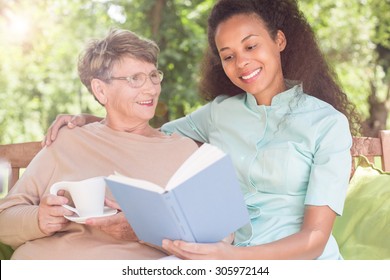 Senior Woman And Caregiver Reading Book In The Garden