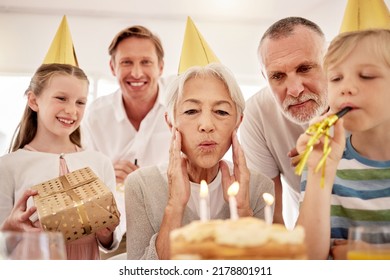 Senior Woman Celebrating A Birthday With Family At Home, Wearing Party Hats And Blowing Whistles. Grandma Looking At Birthday Cake And Being Joyful While Surrounded By Her Grandkids, Husband And Son