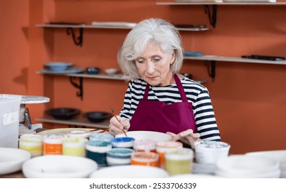 Senior woman ceramist painting new crafted plate with colors and brushes. - Powered by Shutterstock