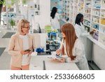 Senior woman chats with a pharmacist at a pharmacy counter, surrounded by pills and drugs. The pharmacist provides advice as they discuss healthcare options