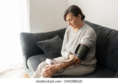 Senior Woman Checking Blood Pressure Level At Home, Older Female Suffering From High Blood Pressure Sitting At A Couch And Using A Pulsometer, Tonometer