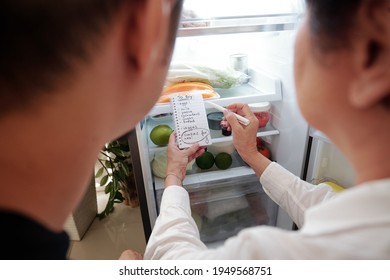 Senior Woman Checking Her Fridge, Writing Shopping List And Asking Adult Son To Buy Her Groceries